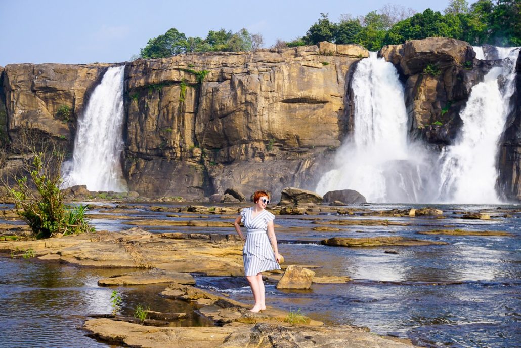 Alice standing on a rock wearing a blue and white striped dress with three cascades from Athirappilly Falls in the background