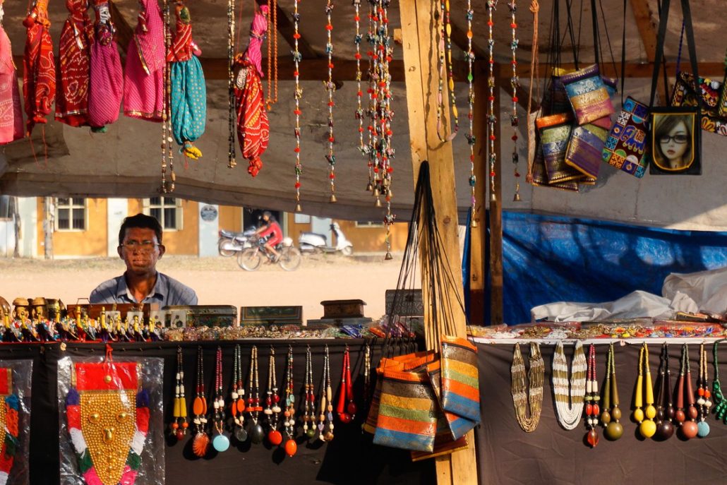 A stall filled with vibrant locally made necklaces and purses hanging in the front of the shop with a man, the shopkeeper, sitting behind the table.