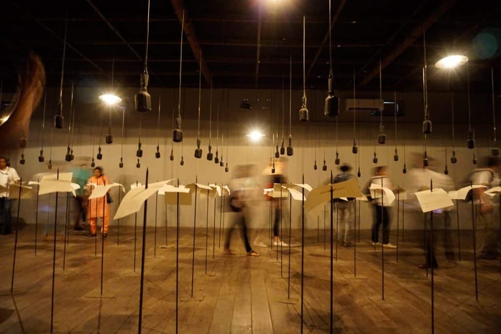 Art installation with 50+ microphones hanging from the ceiling. Under each microphone is a pole punctured through a single yellow slip of paper.