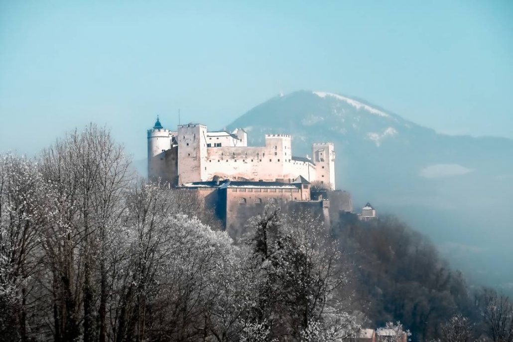 The stark white fortress, Festung Hohensalzburg in Austria with a single mountain behind it in the distance.