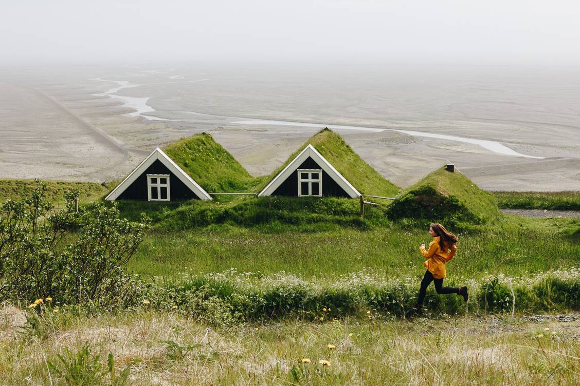 Photo of a girl running across a grassy field with the top of a moss covered house peaking up in the background.