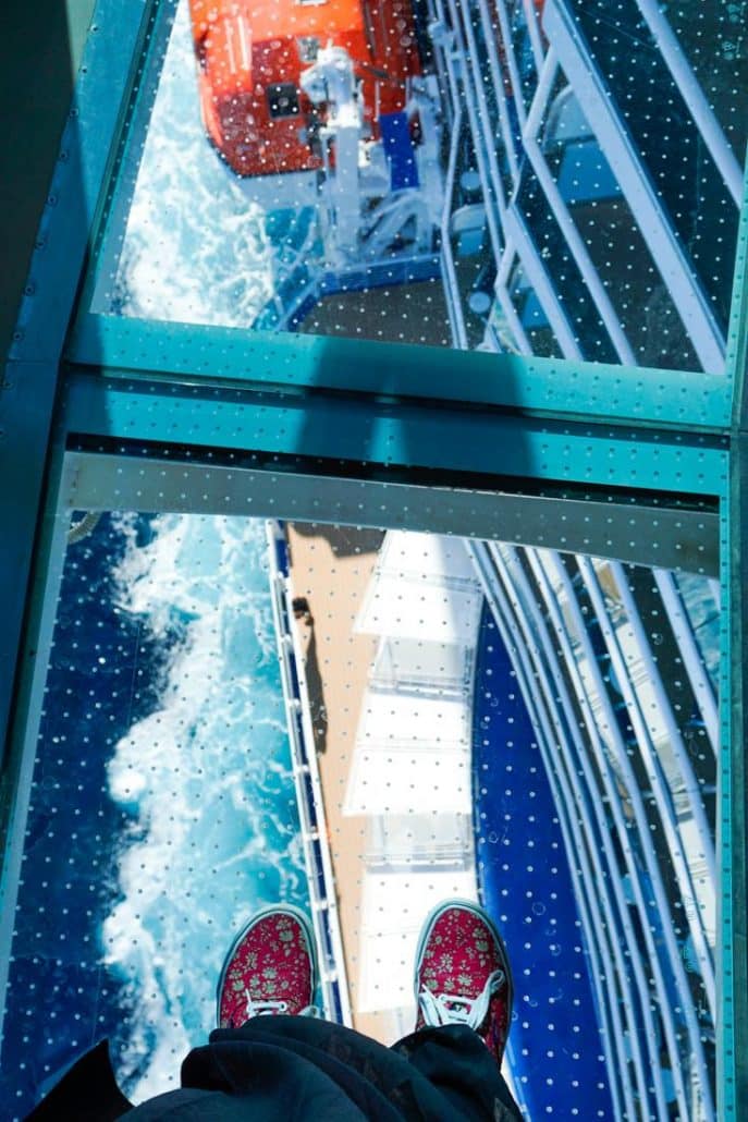 Photo of Alice standing on the glass floor of the seawalk looking down onto the side of the deck and the ocean.